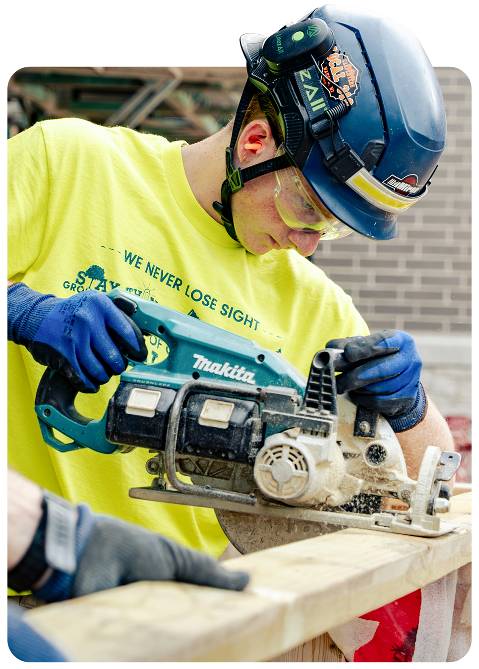A young apprentice in a hard hat and safety glasses cutting lumber with a circular saw