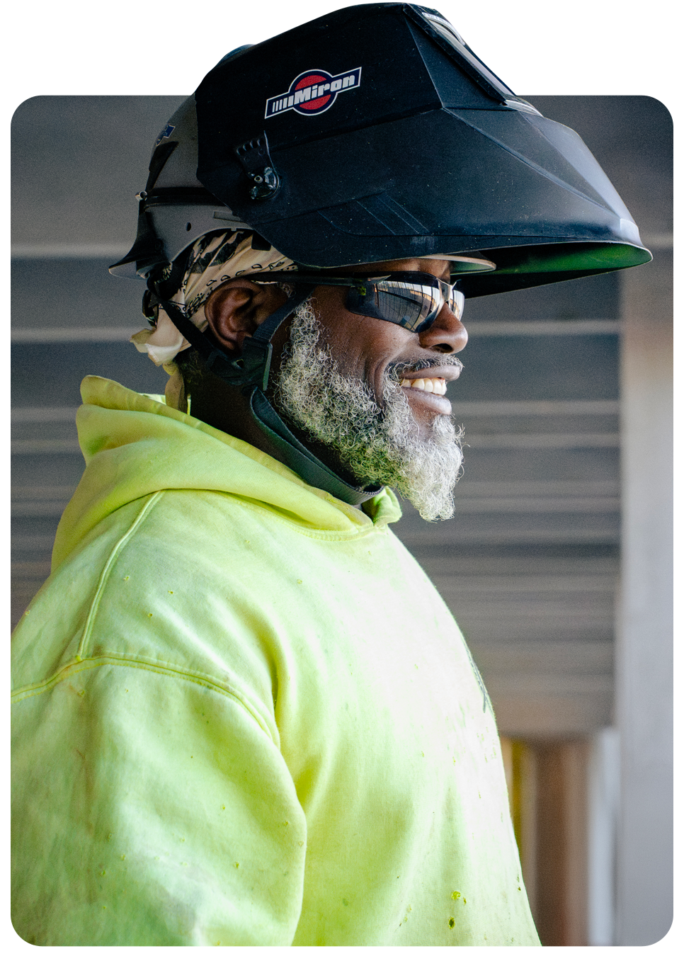 A smiling Miron craftsperson in a welding helmet and high-visibility hoodie