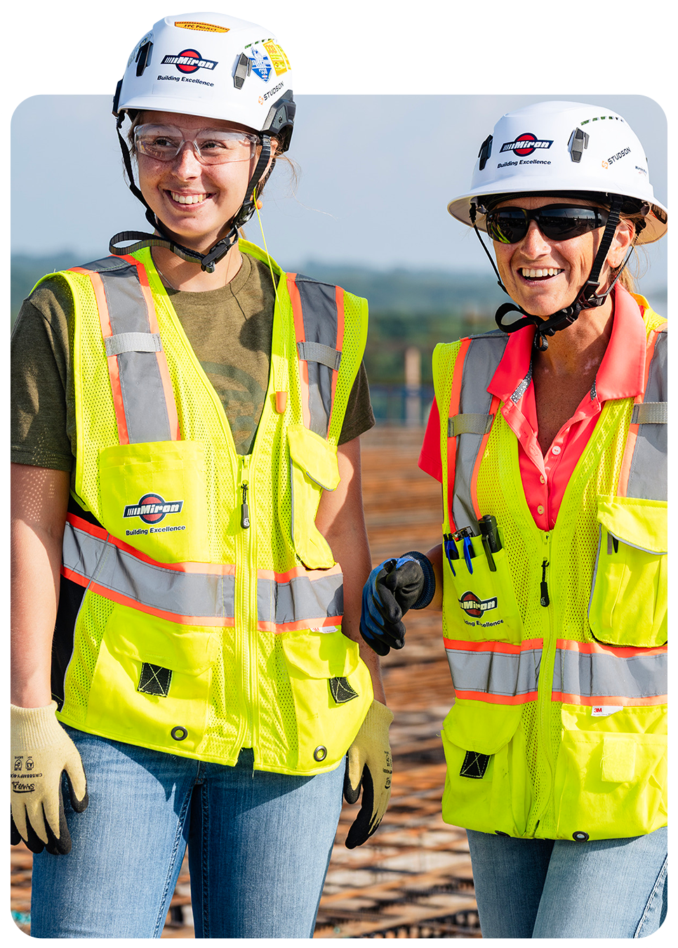 Two smiling interns in hard hats and high-visibility vests on a job site